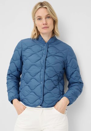 Frau mit blondem Haar, die eine blaue gesteppte Jacke mit Druckknöpfen und weiße Hose trägt, steht mit den Händen in den Taschen vor einem einfarbigen Hintergrund.