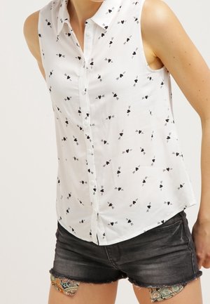 Mouwloos wit overhemd met knopen, met een patroon van zwarte harten en pijlen, gedragen met zwarte denim shorts met geappliceerde stofpatches.