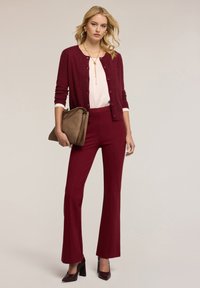 Bordeaux kardigan přes bílou blůzu, doplněný o přiléhavé bordeaux kalhoty a lodičky se špičatou špičkou. Hnědá taška s texturovaným povrchem.