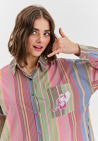 Koszula w paski w różnych kolorach, w tym różowym, zielonym i niebieskim. Posiada dekoracyjną naszywkę i kolorowe guziki.