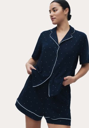 Donna che indossa un pigiama blu navy con maniche corte a bottoni e pantaloncini abbinati con motivo a pois bianchi e rifiniture bianche, mani nelle tasche.