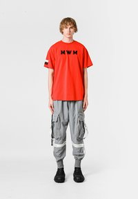 Camiseta de algodón roja con logo "MWM", combinada con pantalones cargo grises con bolsillos y acentos blancos; el modelo lleva puestos zapatos negros texturizados.
