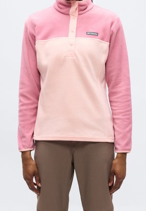 Person iført en tofarvet pink Columbia fleece pullover med lange ærmer og brune bukser mod en ensfarvet baggrund.