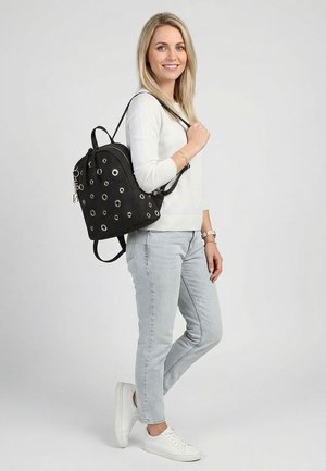 Femme souriante en pull blanc et jean clair, debout avec un sac à dos noir orné d'œillets métalliques sur une épaule, devant un fond uni.