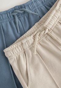 Svetlo béžové a tlmené modré nohavice s šnúrkou v páse, textúrovanou látkou, elastickými pásmi a viditeľnými prednými vreckami vrstvené vedľa seba.