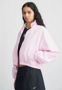 Ροζ κοντός αδιάβροχος με φερμουάρ μπροστά, ελαστικές μανσέτες και λευκό λογότυπο Nike. Ελαφρύ ύφασμα με λεία υφή. Συνδυάζεται με μαύρα ρούχα.