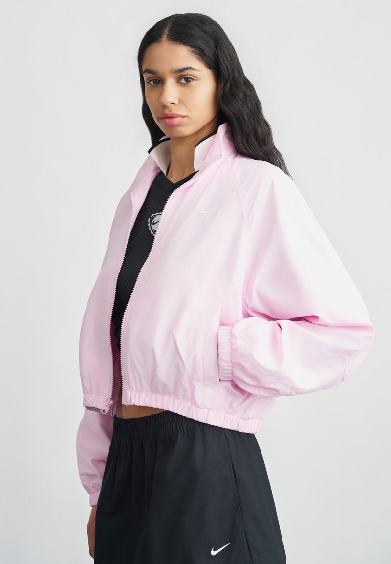 Ροζ κοντός αδιάβροχος με φερμουάρ μπροστά, ελαστικές μανσέτες και λευκό λογότυπο Nike. Ελαφρύ ύφασμα με λεία υφή. Συνδυάζεται με μαύρα ρούχα.