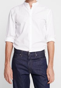 Biała koszula zapinana na guziki z kołnierzem typu band, charakteryzująca się gładką bawełnianą fakturą i podwiniętymi rękawami, noszona z ciemnymi dżinsami.