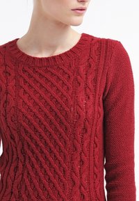 Röd stickad tröja med ett texturerat kabelmönster, ribbad rund hals, och långa ärmar med en slät finish.