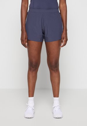 Personne portant un short de sport bleu foncé, des chaussettes blanches et des chaussures de course blanches, debout devant un fond clair uni.