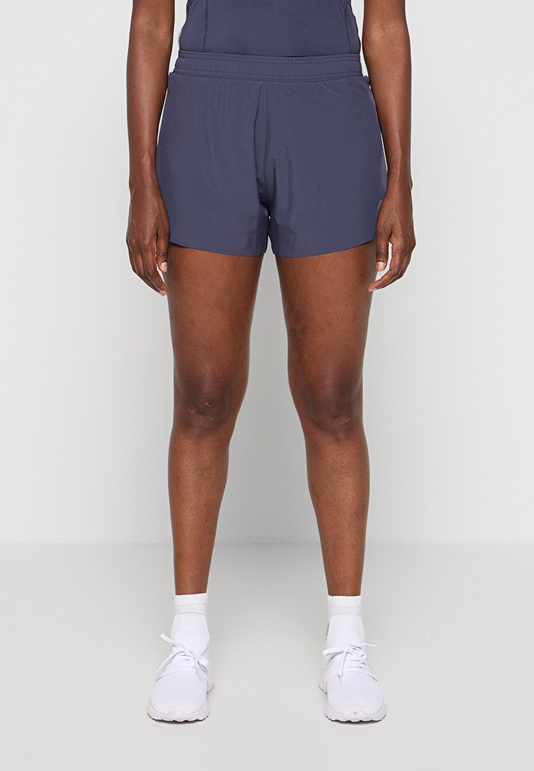 Personne portant un short de sport bleu foncé, des chaussettes blanches et des chaussures de course blanches, debout devant un fond clair uni.