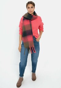 Wollen sjaal in een donkergroen, roze en zwart ruitpatroon. Heeft franjes aan de randen. Gecombineerd met een gestructureerde roze trui en een blauwe jeans.
