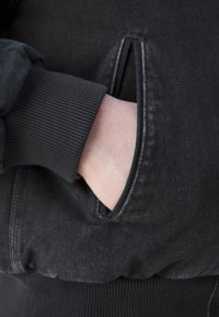 Czarna kurtka z prążkowanymi mankietami, z boczną kieszenią. Materiał wydaje się teksturowany, z kontrastującymi gładkimi akcentami na krawędziach kieszeni.