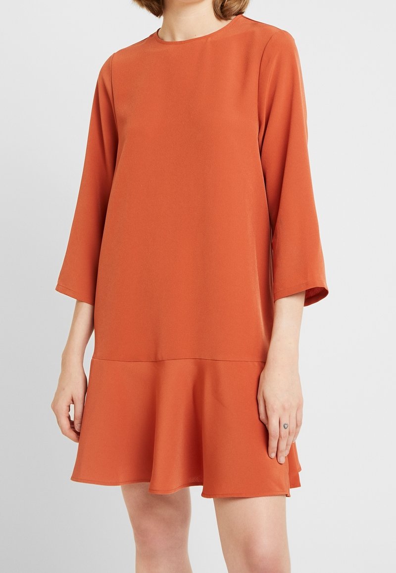 Femme portant une robe ample à manches longues de couleur orange brûlé avec un ourlet évasé, debout devant un fond clair uni.