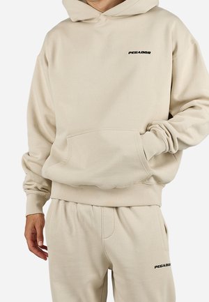 Beigefarbener Pegador-Hoodie mit Fronttasche und passenden Jogginghosen, mit Pegador-Logos auf Brust und Oberschenkel, getragen von einer Person.