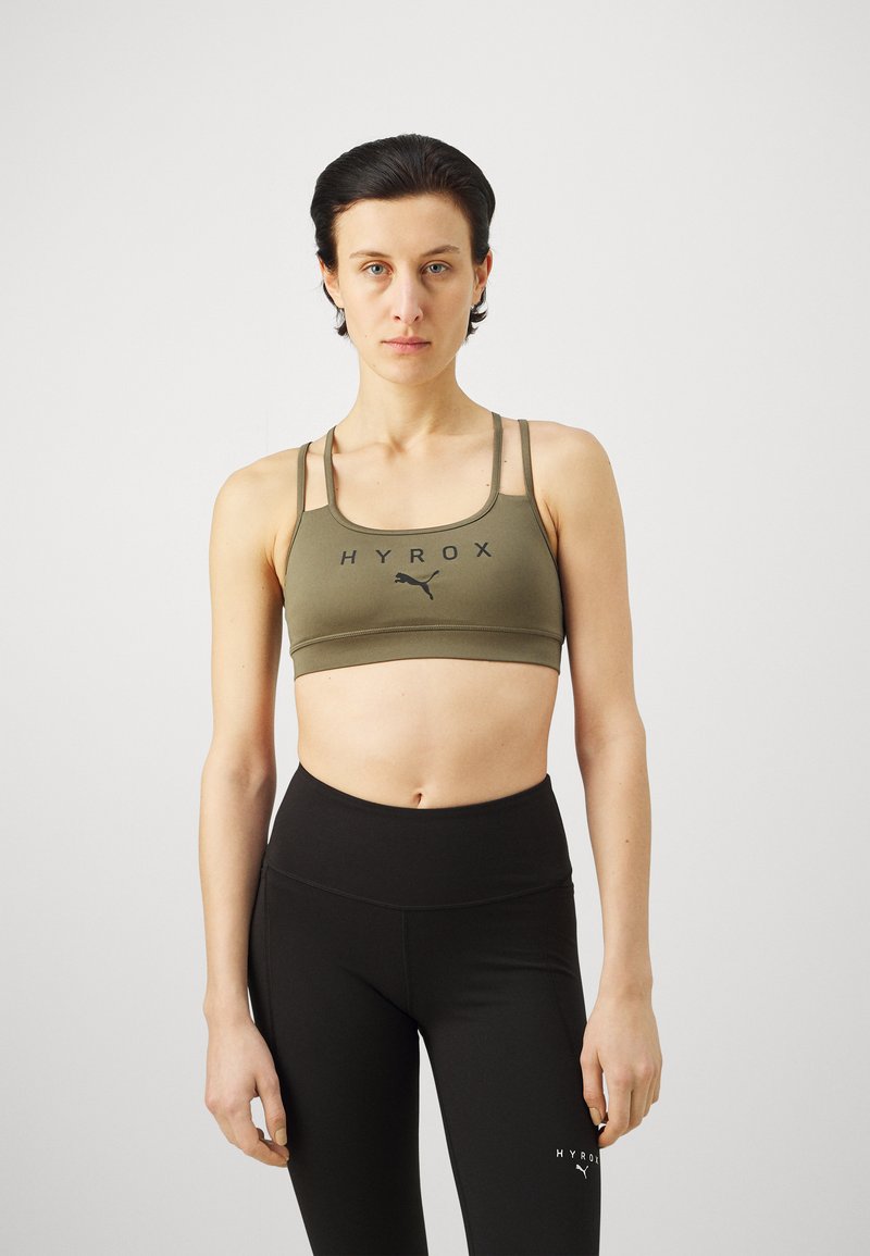 Sujetador deportivo verde oliva con logo negro, con tirantes finos y un diseño acolchado. Combinado con leggings negros de cintura alta.