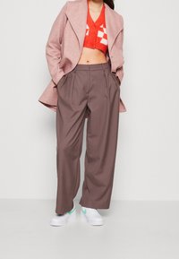 Pantaloni larghi marroni con pieghe, abbinati a un maglione rosso con scacchi bianchi e un cappotto rosa. Completati con scarpe da ginnastica bianche.
