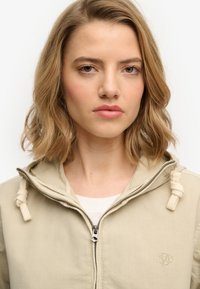 Chaqueta beige con cremallera y capucha con cordón, con una textura suave y logo bordado en el pecho. El modelo muestra una expresión neutral.