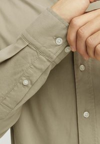 Camisa de manga larga color beige claro con una textura suave, con botones blancos y costuras visibles en los puños y en el placket.