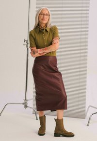 Oliwkowa koszulka polo, bordowa skórzana spódnica oraz brązowe botki za kostkę z żebrowanymi akcentami, stojące na tle w paski.