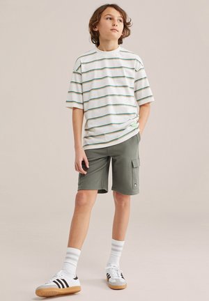 Junge Person steht, trägt ein weißes, gestreiftes T-Shirt, olivgrüne Cargo-Shorts, weiße Socken und weiße Sneaker mit schwarzen Streifen, blickt zur Seite.
