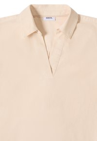 Chemise de couleur crème avec un col et un décolleté en forme de V ouvert, fabriquée dans un tissu texturé, étiquetée "CECIL" à l'intérieur du col.