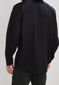 Man som bär en långärmad svart skjorta och mörka cargobyxor, sedd bakifrån mot en enkel ljus bakgrund.