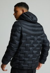 Mustmust must jacket, millekiliste mustriga, paksu kapuutsiga ja elastsete mansettidega. Valmistatud kergest, läikivast materjalist, millel on sile tekstuur.