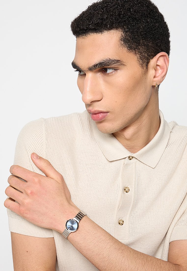 Zilveren horloge met een ronde, blauwe wijzerplaat en metalen band; gedragen om een lichtgekleurde arm. Crème-kleurig, strak gebreid poloshirt.