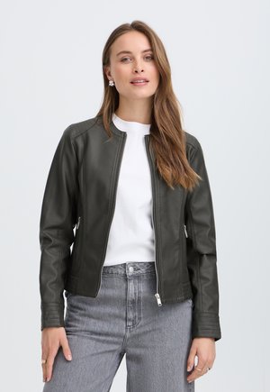 Femme aux longs cheveux bruns portant une veste en cuir noire, une chemise blanche et un jean gris, debout devant un fond uni clair.