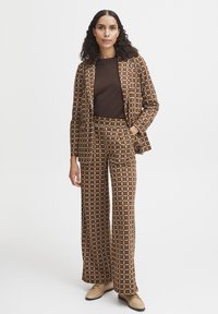 Costume à carreaux marron avec un blazer à motifs et un pantalon large. Le tissu présente des carrés marron, crème et bleu. Haut marron et chaussures claires.