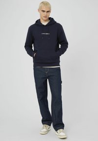 Sweat à capuche bleu marine avec poche kangourou, capuche à cordon de serrage et détails brodés. Associé à un jean sombre et des baskets beiges.