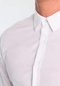 Bílá bavlněná košile na knoflíky s klasickým límečkem, která má hladkou texturu a skrytou légu knoflíků.