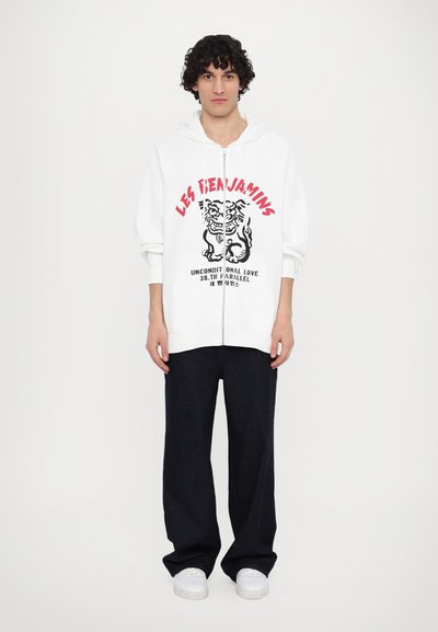 Λευκή φούτερ ζακέτα με φερμουάρ και μαύρη και κόκκινη γραφική εκτύπωση που απεικονίζει μια τίγρη και κείμενο, συνδυασμένη με σκούρο τζιν παντελόνι και άσπρα αθλητικά παπούτσια.