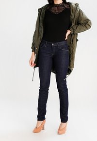 Olivově zelená parka se stahovací šňůrkou, černý žebrovaný top s krajkovým detailem, tmavě modré úzké džíny a nahé boty na vysokém podpatku.