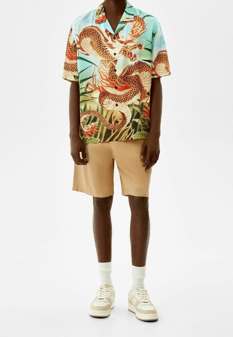 Krátký rukáv košile s barevným potiskem draka na modrém pozadí, doplněná béžovými šortkami a bílými teniskami s krémovými akcenty.