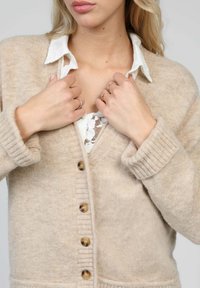 Cardigan en laine beige avec des poignets côtelés et une fermeture centrale à boutons. Superposé à une chemise blanche à col avec un détail en dentelle.