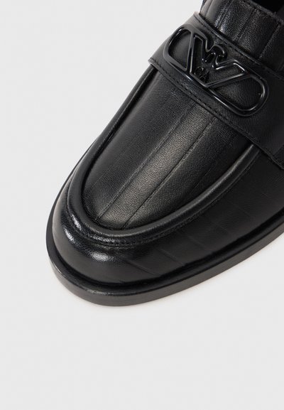 Musta nahkainen loafer, jossa on sileä pinta, teksturoidut yksityiskohdat ja suuri musta logo-nauha. Ominaisuuksia ovat pyöristetty kärki ja litteä pohja.
