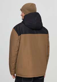 Brązowa kurtka puchowa z czarnym kapturem, charakteryzująca się gładką fakturą i luźnym krojem. Na głowie brązowy, żebrowany beanie.