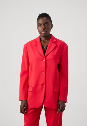 Blazer rouge en tissu lisse, design à un bouton, avec des revers crantés, trois boutons et poches avant. Stylisé avec un haut noir en dessous.