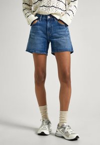 Denim kratke hlače v srednji modri barvi s ravnim krojem, z sprednjimi žepi, zankami za pas in čisto robom. Upoštevane so s sivimi športnimi čevlji in svetlimi nogavicami.