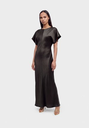 Femme portant une robe longue en satin noir à manches courtes et des sandales dorées à talons hauts à brides, debout sur un fond blanc uni.