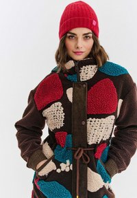 Žena oblečená v červenom čiapke a farebnej patchworkovej fleecovej bunde, s rukami v vreckách, pózujúca na bielom pozadí.