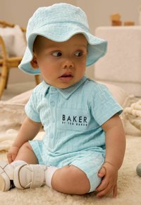 Mono corto de manga corta azul claro con un patrón texturizado y detalles de botones, que presenta un logo de "BAKER" y un sombrero de cubo a juego. Calzado blanco.