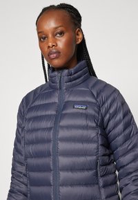 Tamsiai mėlynas pūkinis švarkas su aukštu apykakle, siuvinėta struktūra, priekinis užtrauktukas ir mažas prekės ženklas ant kairės krūtinės.