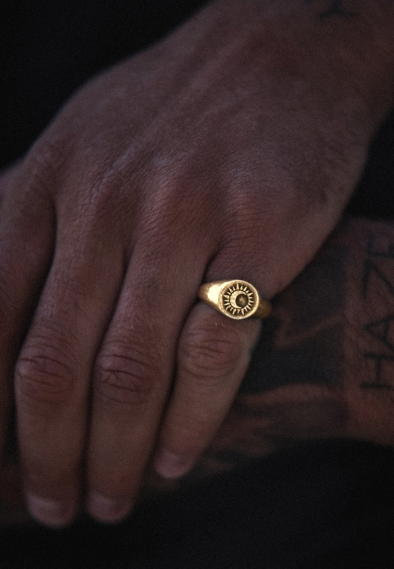 Kuldne sõrmus päikesepaiste kujundiga, millel on sile, poleeritud pind ja ümar kuju. Käsi näitab tekstuuriga nahka.