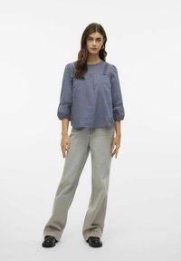 Modrá texturovaná blůza s vycpanými rukávy, čtveratým výstřihem a jemnými puntíky, spárovaná s šedými kalhotami s širokými nohavicemi a černými botami.