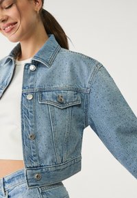Svijetloplava denim jakna skraćenog kroja s srebrnim zakovicama, zatvaranjem na gumbe, dva prednja džepa i ovratnikom. Glatka tekstura, blago prilagođen dizajn.