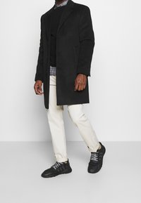 Schwarzer Mantel, grauer Pullover, kariertes Hemd sichtbar, cremefarbene Hose und schwarze Sportschuhe mit grauen Akzenten und Schnürsenkeln. Glattes, strukturiertes Design.