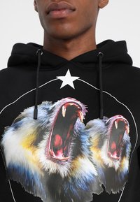 Hoodie noir avec un motif vivant de deux lions rugissants, rehaussé d'une étoile blanche et d'un design de lignes courbes. Texture de tissu douce.